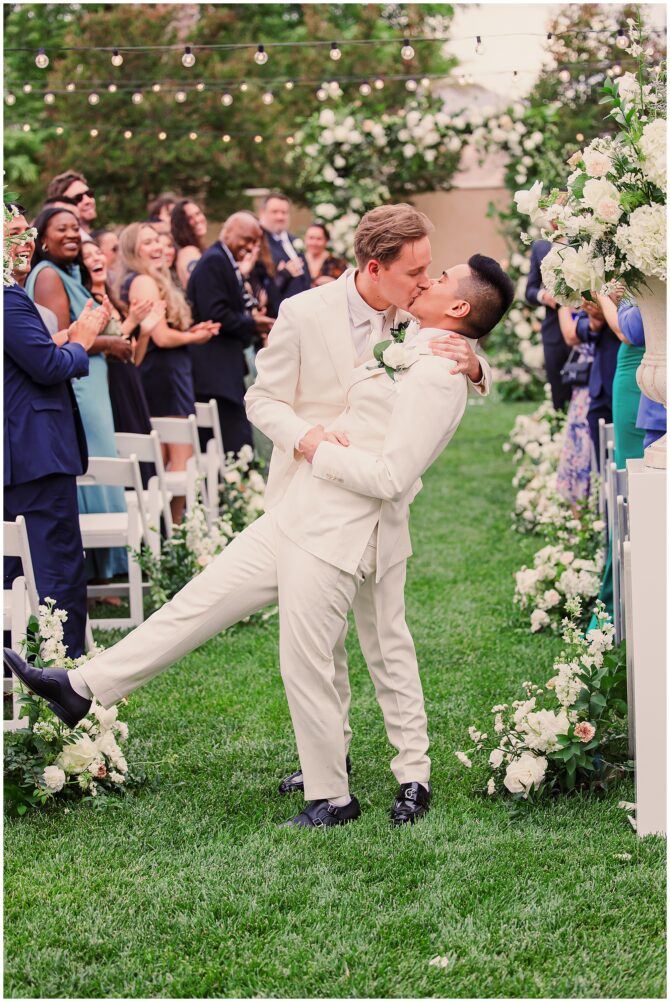 Two grooms kissing at outdoor wedding ceremony.