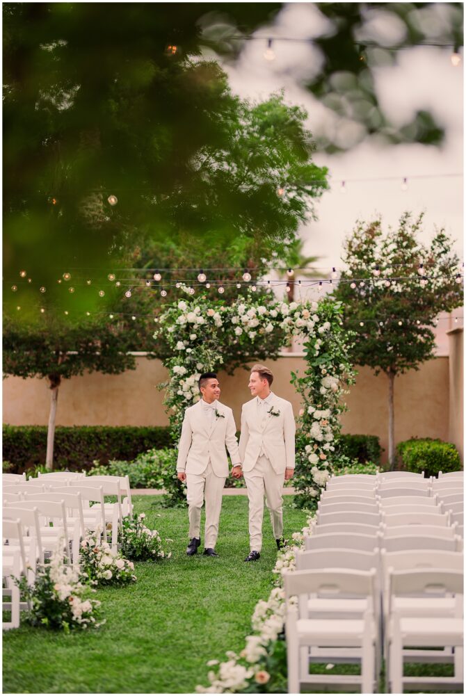 Two grooms walking down outdoor wedding aisle.