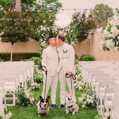 Grooms kiss with dogs at outdoor wedding ceremony.
