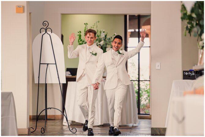 Couple in white suits celebrating at wedding entrance.
